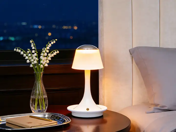 Illuminated white table lamp on a round bedside table beside a glass vase and a tray