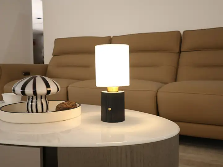 Table lamp with a white cylindrical shade and dark base on a coffee table in front of a tan sofa